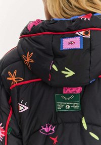 Černá prošívaná bunda s barevnými abstraktními vzory, červenými akcenty a několika štítky s různými motivy a textem na zádech.