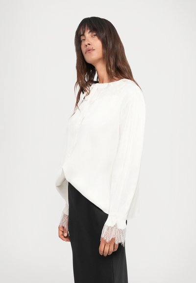 Weißes Blusenshirt mit rundem Halsausschnitt, Spitzenbesatz an den langen Ärmeln und lockerer Passform, kombiniert mit einem schwarzen Rock. Glatte Stofftextur.