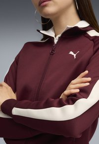 Bordová zipsová bunda s bielymi akcentmi, s vysokým golierom a logom Puma. Textúra je hladká s lemovanými manžetami a detailmi na leme.