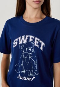 Femme portant un t-shirt bleu avec une illustration de teddy bear blanc et le texte "SWEET dreams" sur le devant.