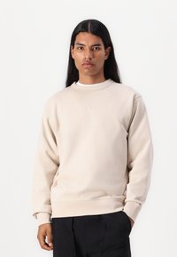Sweatshirt bege claro com decote redondo, punhos canelados e uma textura suave. Apresenta um discreto detalhe de costura diagonal na zona do pescoço.