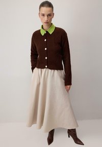 Brūns adīts kardigans ar zaļu apkakli, rotāts ar dimanta rakstu un baltām pogām, saskaņots ar gaismas krēma plūstošu svārku un brūniem asiem zābakiem.