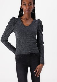 Black glittery long-sleeve top with a V-neck, featuring puffed shoulders and a fitted design, paired with black jeans adorned with studs.