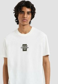 Camiseta de algodón blanca con un gráfico rectangular negro en el centro que contiene el texto "Armani Exchange" y "1991". Diseño de cuello redondo y mangas cortas.