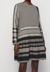 Schwarz-weiß gemustertes Kleid mit langen Ärmeln, das geometrische Designs und einen abgestuften Rock zeigt. Hergestellt aus leichtem Stoff, mit rundem Ausschnitt.