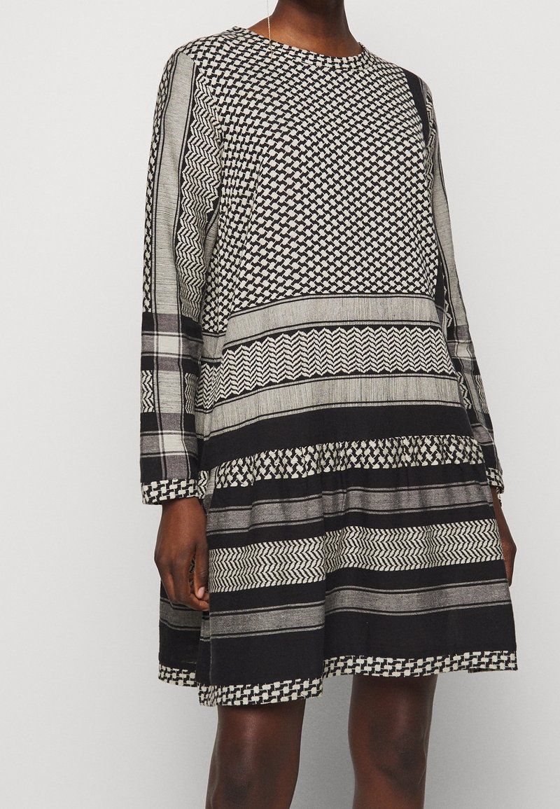 Schwarz-weiß gemustertes Kleid mit langen Ärmeln, das geometrische Designs und einen abgestuften Rock zeigt. Hergestellt aus leichtem Stoff, mit rundem Ausschnitt.