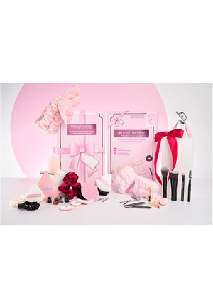 Calendario de adviento de belleza Brush Works con brochas de maquillaje, esponjas, clips para el cabello, scrunchies, rizador de pestañas y diademas sobre un fondo rosa y blanco.