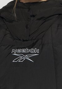 Veste noire Reebok avec logo et emblème sur la poitrine, équipée de deux boutons-pression noirs près du col.