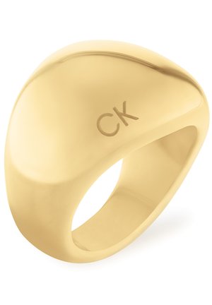 Anillo dorado con forma suave y redondeada que presenta el logotipo de CK. La superficie está pulida, reflejando la luz con una textura elegante.