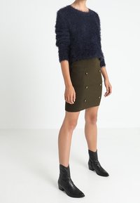 Marineblauer, flauschiger Strickpullover, olivgrüner, figurbetonter Rock mit goldenen Knöpfen und schwarze spitze Stiefeletten mit geprägten Details.