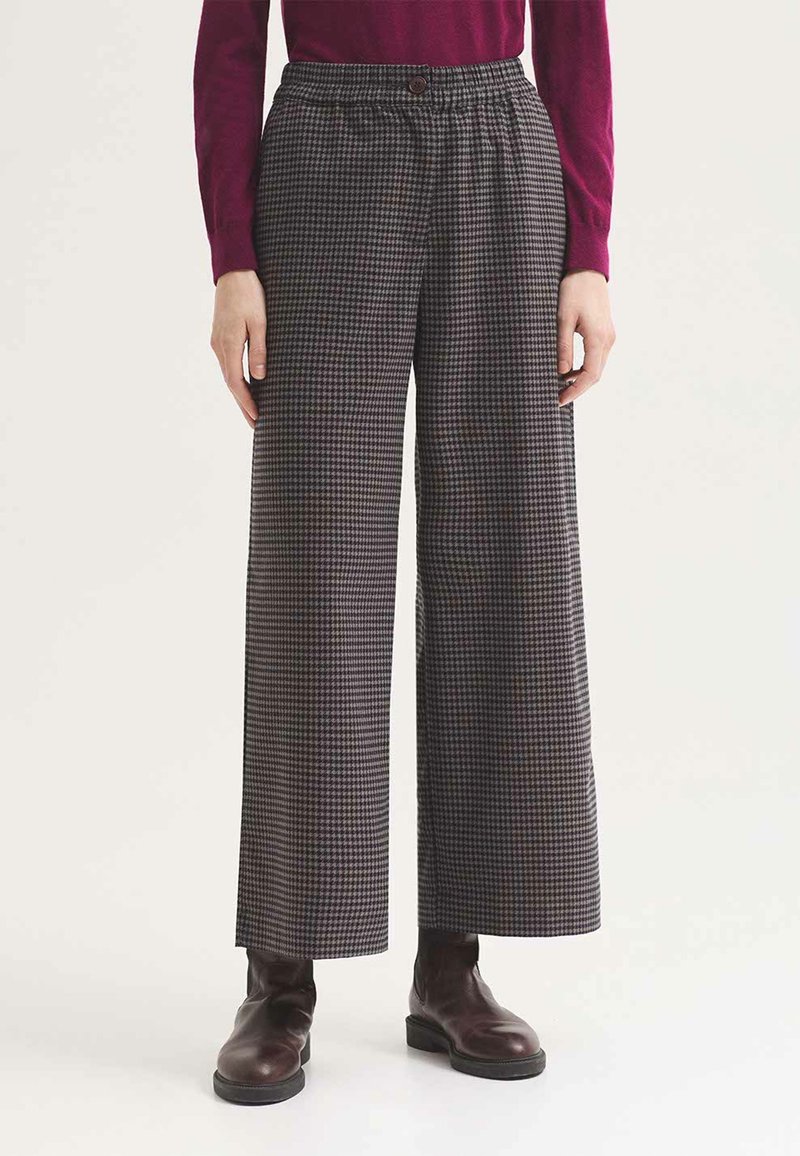 Personne portant un pantalon taille haute à jambes larges à motif pied-de-poule avec fermeture par boutons, un haut bordeaux à manches longues et des bottines marron foncé.