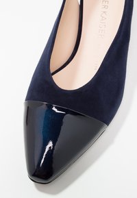 Escarpin en daim bleu marine avec un bout pointu en cuir verni brillant. Présente un devant échancré et une finition texturée sur la partie supérieure.