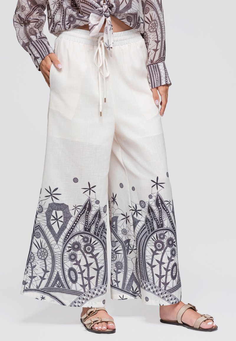 Pantalon blanc à jambes larges avec imprimé floral géométrique noir à l'ourlet, taille élastique avec cordon, associé à des sandales beige et une chemise imprimée nouée.
