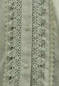 Κοντινό πλάνο στρογγυλών κουμπιών σε ανοιχτό πράσινο ύφασμα με περίτεχνη δαντέλα που διαθέτει λουλουδένια μοτίβα και κυματιστές άκρες.