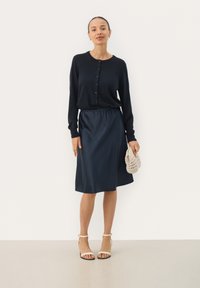 Mörkblå knälång kjol med resår i midjan, ihop med en svart knapp-up cardigan och vita sandaler med remmar. Hållen beige väska.
