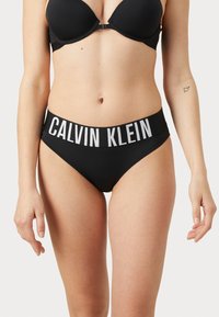 Svart bikinitruse med bred linning merket "CALVIN KLEIN" i hvitt. Glatt stoff, moderat dekning og sømløse kanter.