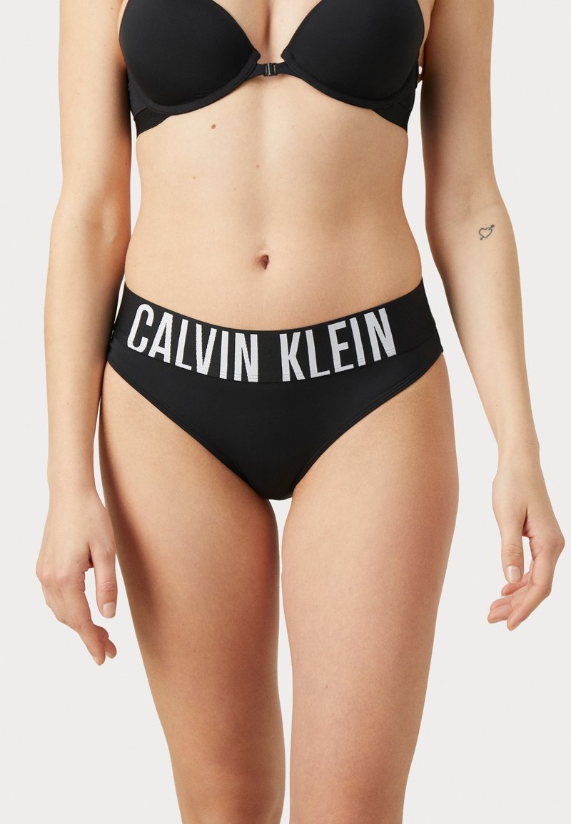 Svart bikinitruse med bred linning merket "CALVIN KLEIN" i hvitt. Glatt stoff, moderat dekning og sømløse kanter.