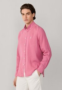 Jeune homme aux cheveux bruns portant une chemise rose à boutons et un pantalon blanc, debout avec une main dans la poche devant un fond uni.