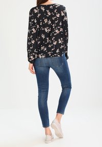 Blouse noire à manches longues et à coupe ample, ornée de fleurs roses et blanches ; associée à un jean skinny bleu et à des chaussures gris clair.