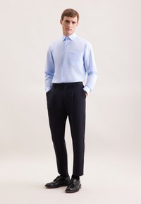 Camicia azzurro chiaro con taschino, abbinata a pantaloni neri slim e scarpe nere, su uno sfondo neutro.