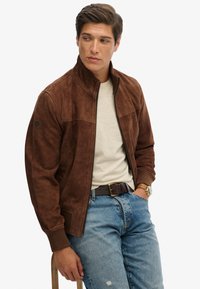 Giacca bomber in suede marrone con colletto alto, chiusura con zip frontale e polsini a costine. Abbinata a una camicia chiara e jeans blu strappati.