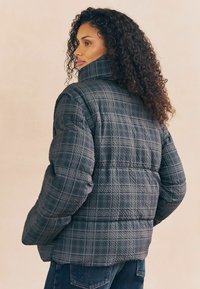 Vrouw met krullend haar in een grijze geruite pufferjack en blauwe jeans, zijwaarts staand met handen in de zakken.