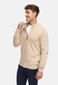 Lichtbeige ritscardigan, gemaakt van zacht materiaal, met een geribbelde kraag en manchetten, voorzien van een klein bruin logo-embleem op de zoom.