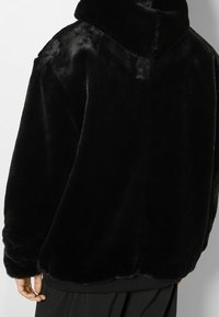 Giacca in pelliccia sintetica nera con colletto alto, vestibilità oversize e texture morbida, caratterizzata da un orlo arrotondato e polsini elastici.