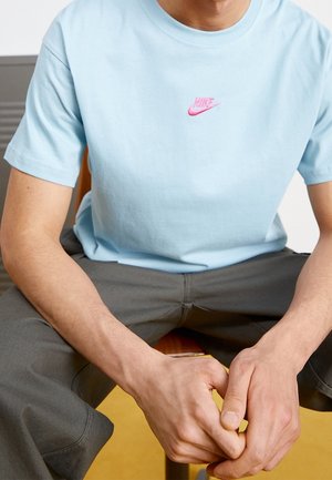 Mężczyzna siedzi na krześle, ubrany w jasno niebieską koszulkę Nike i ciemnoszare spodnie, ręce splecione między nogami.