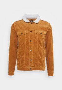 Veste en velours côtelé couleur ambre avec un col en fausse fourrure blanche, deux poches poitrine et une fermeture par boutons. Tissu texturé à côtes verticales.