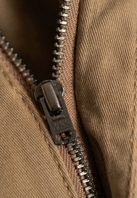 Tissu marron avec une vue rapprochée d'une fermeture éclair YKK, présentant un curseur et des dents métalliques, mettant en valeur les détails de la texture et des coutures.