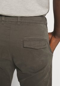 Calças verde-escuras com uma textura suave, apresentando um bolso traseiro e passantes para cinto. A cintura é plana e o tecido parece ligeiramente escovado.