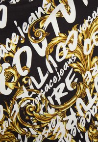 Černá látka s výrazným bílým textem a zdobnými zlatými barokními vzory. Text obsahuje "Versace Jeans" v různých velikostech a stylech.