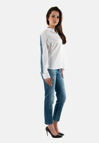 Camisa blanca de botones con un ribete azul estampado en las mangas, combinada con jeans azul claro y tacones negros de punta. Textura de tela suave.