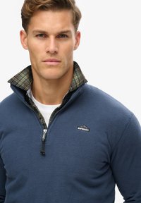 Sudadera de polar azul con cuello con cremallera, que presenta un forro de tartán a cuadros y un parche de logo. Textura suave con un ajuste relajado.