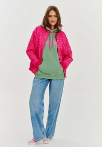 Ružová prešívaná bunda cez zelenú mikinu s textom "bonjour", v kombinácii s voľnými modrými džínsami a ružovými teniskami.
