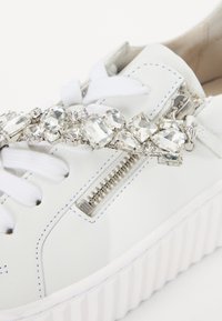 Hvide lædersneakers med en struktureret hvid sål, der har en krystaludsmykning langs siden og en sølv lynlåsdetalje.