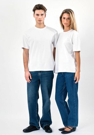 Ung mand og kvinde står side om side iført ensfarvede hvide t-shirts og blå jeans mod en hvid baggrund.