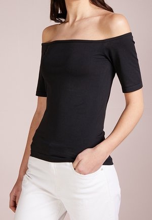 Femme portant un haut noir ajusté, épaules dénudées avec manches courtes et un pantalon blanc, une main dans la poche, sur fond neutre.