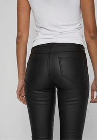 Pantalon noir en similicuir avec une texture lisse, coupe ajustée et coutures visibles. Comprend deux poches arrière et une taille mi-haute.