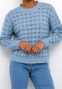 Lichtblauwe gebreide trui met een gestructureerd open patroon, geribde ronde hals en manchetten. Gecombineerd met lichtblauwe jeans, handen voor elkaar.
