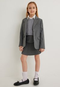 Grijze schoolblazer met zilveren knopen, een grijze gebreide trui, een geplooide grijze rok, een wit shirt, witte sokken en zwarte platte schoenen.