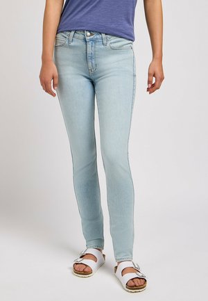 Jean skinny bleu clair en denim, doté d'un design classique à cinq poches et d'une fermeture à bouton, associé à des sandales blanches.