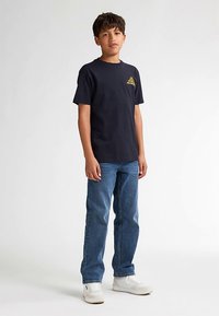 Laivastonsininen puuvillainen T-paita, jossa on keltainen logo rinnassa, yhdistetty farkkuihin ja valkoisiin tennareihin neutraalilla taustalla.