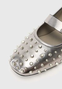 Metalli hõbedane king, millel on ümar nina, millel on erineva suurusega studded pind ja Mary Jane rihm ülevalt.