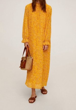 Personne portant une longue robe jaune fleurie à manches longues, tenant un sac en paille avec des lanières en cuir marron, et portant des sandales marron.