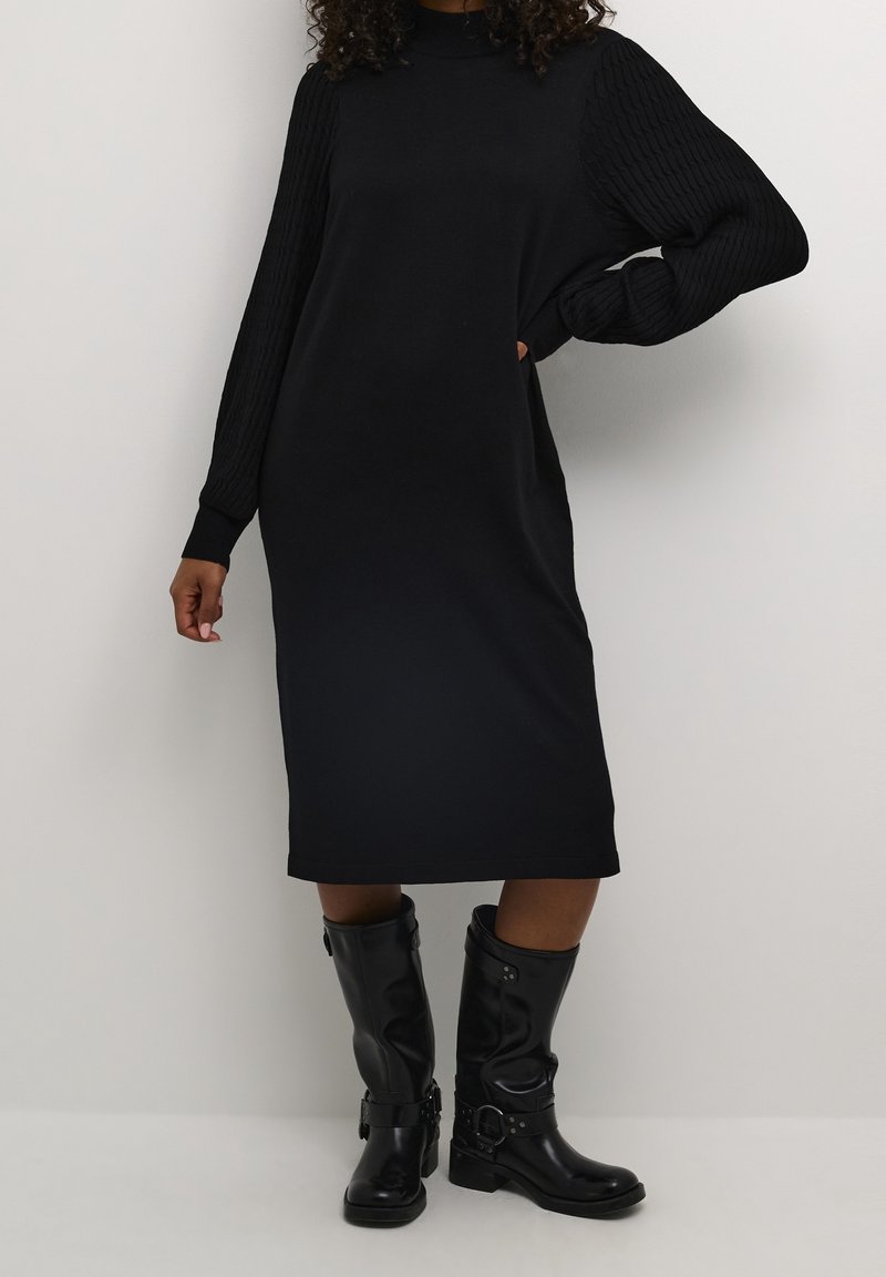 Personne portant une longue robe noire avec des manches texturées et des bottes en cuir noires mi-mollet, debout devant un fond blanc.
