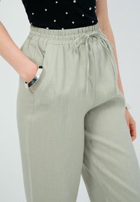 Světle šedé kalhoty s elastickým pasem a šňůrkou, které mají boční kapsy a jsou vyrobené z měkké, strukturované tkaniny. Bez viditelných vzorů nebo kovových komponentů.