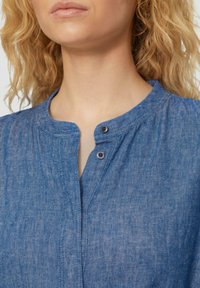 Blaues Jeanshemd mit rundem Ausschnitt, zwei schwarzen Druckknöpfen und einer leichten Struktur. Der Stoff wirkt weich und leicht.