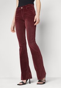 Pantalones acampanados de pana burdeos con una textura suave, bolsillos frontales y cierre de un solo botón. Combinados con tacones negros de tiras.
